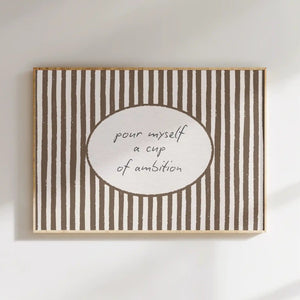 A rectangular framed wall art print with vertical brown and white stripes and a white oval center containing the handwritten phrase 'pour myself a cup of ambition,' shown hanging on a white wall with soft shadows.
